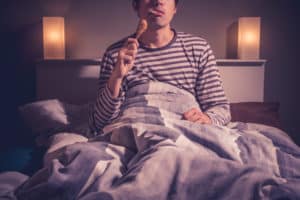 Young man is sitting in bed at night and eating a chicken drumstick