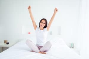 young caucasian woman sitting on bed looking happy and arms in the air