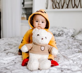 Child in raincoat sitting on bed holding teddy bear