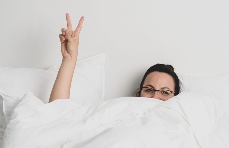 image of woman in bed with white sheets and throwing a peace sign